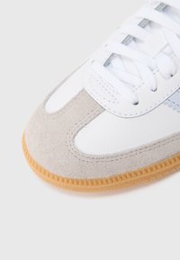 Λευκό αθλητικό παπούτσι με γκρι σουέντ λεπτομέρειες και ανοιχτομπλε ραφές. Διαθέτει υφασμένο λάστιχο σε καφέ απόχρωση και παραδοσιακό σύστημα κορδονιών.