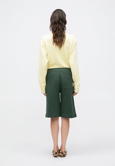 Κίτρινο φούτερ με χαμηλωμένους ώμους συνδυασμένο με πράσινα σορτς μέχρι το γόνατο, με πίσω τσέπη και ελαστική μέση. Παπούτσια με λεοπάρ σχέδιο.