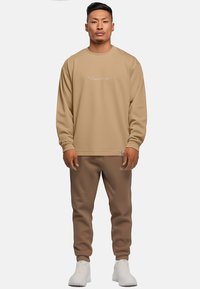 Felpa beige con logo bianco, abbinata a pantaloni jogger beige coordinati, caratterizzati da una vestibilità rilassata e un tessuto testurizzato. Sneakers bianche completano l'outfit.
