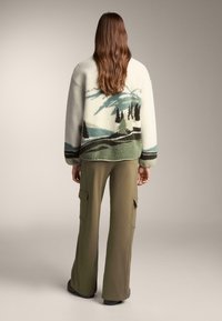 Kurtka polarowa z krajobrazowym wzorem w kolorach zielonym i czarnym, zestawiona z oliwkowymi spodniami typu cargo. Posiada luźny krój i teksturowaną tkaninę.