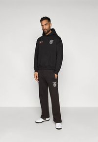 Muž oblečený v čiernej mikine a nohaviciach so športovými logami, stojí s jednou rukou v vrecku a má na sebe biele tenisky na jednoduchom pozadí.