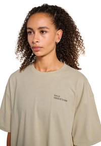Jeune femme aux cheveux bouclés portant un t-shirt beige oversize avec un petit texte noir "HALO 196819721981" sur la poitrine, regardant légèrement à gauche.