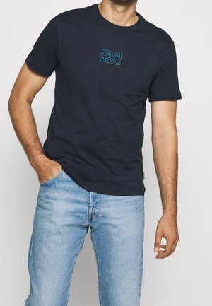 Camiseta de algodón azul marino de manga corta y cuello redondo. Presenta un logotipo rectangular azul con "CALVIN KLEIN" en el centro.