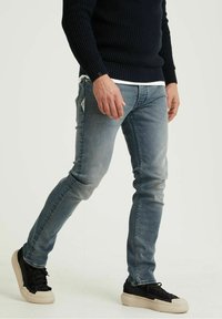 Jeans azzurri chiari con vestibilità slim, caratterizzati da leggere strappi sulla parte anteriore, abbinati a sneakers nere e a un maglione scuro con texture.