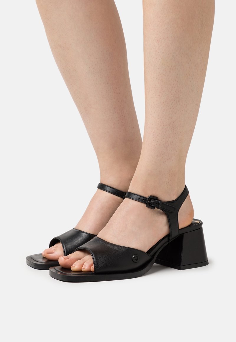Sandales en cuir noir avec un bout ouvert carré, une bride de cheville avec boucle, et un talon bloc épais. Texture lisse et détails métalliques minimaux.