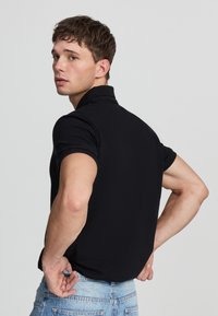 Polo shirt nero a maniche corte, design aderente, texture liscia, con colletto classico. Il modello indossa jeans di denim blu chiaro, in una postura rivolta.