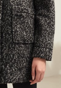 Czarny i szary puchowy płaszcz z wełny o luźnym kroju, z boczną kieszenią i lekko podwiniętymi mankietami.