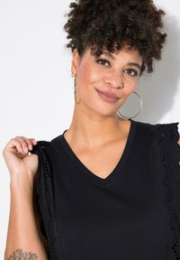 Top nero a scollo a V con dettagli in pizzo sulle spalle, caratterizzato da una vestibilità morbida e una texture morbida. Il modello posa, tenendo il tessuto con un leggero sorriso.
