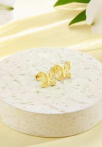 Guldörhängen i form av fjärilar med intrikata utskurna mönster, som vilar på en texturerad ljus sten cirkulär display, mot en blek gul bakgrund.
