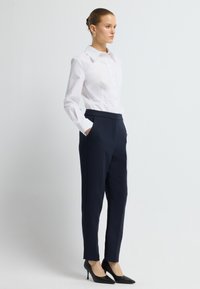 Bílá košile s límečkem, kombinovaná s námořnickými kalhotami na míru. Modelka má na sobě černé texturované lodičky na vysokém podpatku. Jednoduchý, moderní design.