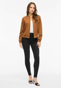 Jachetă bomber maro cu fermoar în față, material ușor, manșete rib, și două buzunare cu fermoar, purtată peste un top alb și blugi negri.