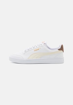 Baskets blanches basses avec bande beige Puma, lacets blancs et languette de talon marron sur un fond clair uni.