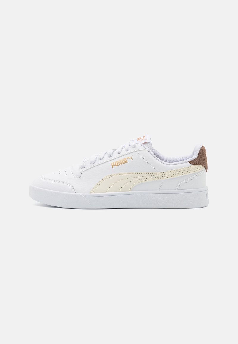 Baskets blanches basses avec bande beige Puma, lacets blancs et languette de talon marron sur un fond clair uni.
