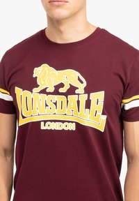 Burgundių spalvos medvilnės marškinėliai su geltonu liūto grafiku ir „Lonsdale London“ tekstu, su baltais ir geltonais dryžiais ant rankovių.