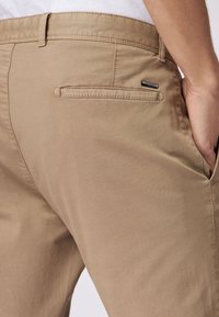 Pantalones de algodón beige con una textura suave, presentan un bolsillo trasero con un logo sutil y un corte ajustado.