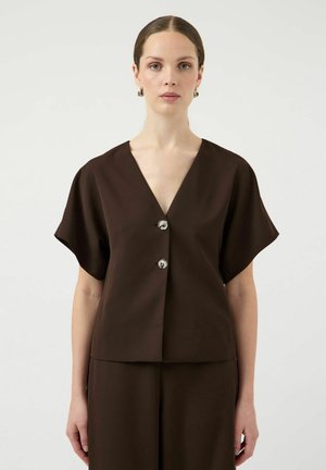 Kobieta z zaczesanymi do tyłu włosami, ubrana w ciemnobrązową, krótko rękawkową, zapinaną na guziki bluzkę oraz pasujące spodnie, stojąca na tle jednolitego jasnego tła.