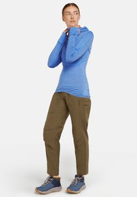Sudadera de cremallera azul claro de manga larga con un diseño ajustado. Pantalones verde oliva ajustados con bolsillos laterales de cremallera. Zapatos azules con acentos grises.