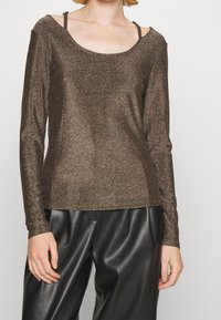 Glänsande brun långärmad topp med rundad halsringning och öppna axelremmar, matchad med svarta läderbyxor.