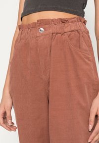 Hnědé kordové kalhoty s nařaseným pasem, stříbrným drukem a bočními kapsami; látka má vertikální proužkování a jemnou strukturu.