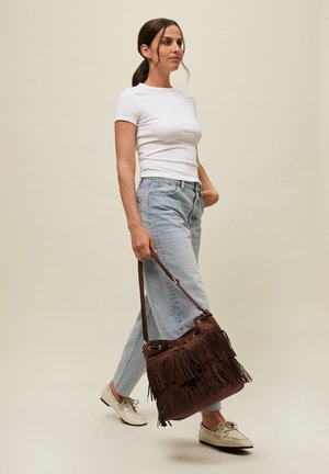 Woman wearing white t-shirt and light blue jeans, walking barefoot while holding a large brown fringed handbag by its strap.