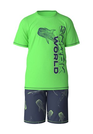 T-shirt verde con grafica di squalo blu navy e testo "SHARK WORLD"; abbinata a pantaloni corti blu navy con stampe di squali chiare.