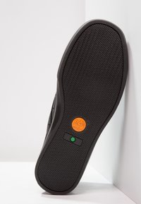 Musta kumipohja, jossa on teksturoitu siksak-kuvio. Siinä on oranssi pyöreä logo ja vihreä suorakulmainen yksityiskohta.