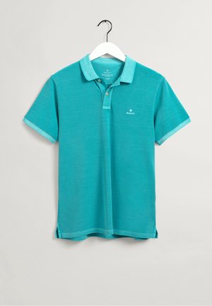 Turkis kortærmet polo med krave og knapper, hængende på en hvid bøjle mod en ensfarvet lys baggrund.