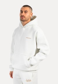 Haut à capuche blanc avec poche kangourou, poignets côtelés et logo brodé en beige clair sur la poitrine.