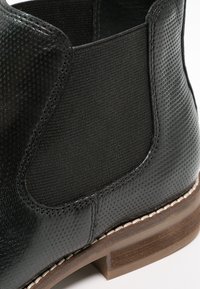 Stivale alla caviglia in pelle nera con superficie texture, pannello laterale elasticizzato e tacco in legno marrone. Dettaglio di cucitura lungo la suola.