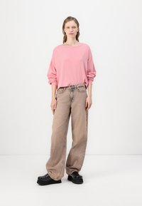 Világos rózsaszín, oversized pulóver, világosbarna, bő szárú farmernadrág, fekete platform cipő, fehér háttér előtt, semleges fényben állva.