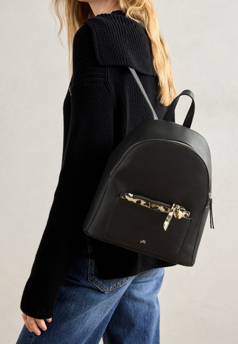Personne portant un pull en maille noir et un jean bleu, portant un sac à dos en cuir noir avec une poche zippée à imprimé léopard à l'avant.