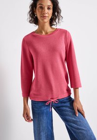 Frau trägt einen rosa Strickpullover mit Dreiviertelärmeln und einem Kordelzugsaum, kombiniert mit blauen Jeans, und steht vor einem weißen Hintergrund.