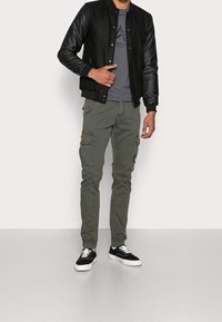 Man står iklädd en svart varsity-jacka, grå grafisk t-shirt, olivgröna cargobyxor med lappar och svarta sneakers med vita sulor.