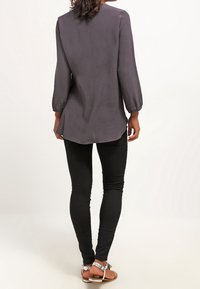 Langärmlige, dunkelgraue Bluse mit dezentem Schimmer, abgerundetem Saum und lockerem Schnitt, kombiniert mit schwarzen Skinny-Jeans und braunen Sandalen.