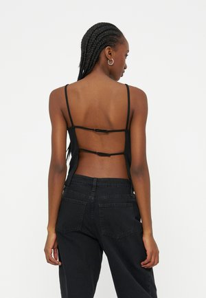 Γυναίκα με κοτσίδες, φοράει μαύρο τοπ με λουράκια και ανοιχτή πλάτη και μαύρο τζιν με ψηλή μέση, απεικονίζεται από πίσω με φόντο ένα απλό φόντο.