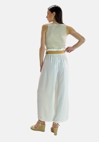 Lys beige udenærmet top, hvide bukser med brede ben og rynket talje, samt brune wedge-sandaler med ankelbånd. Glat tekstur, afslappet design.
