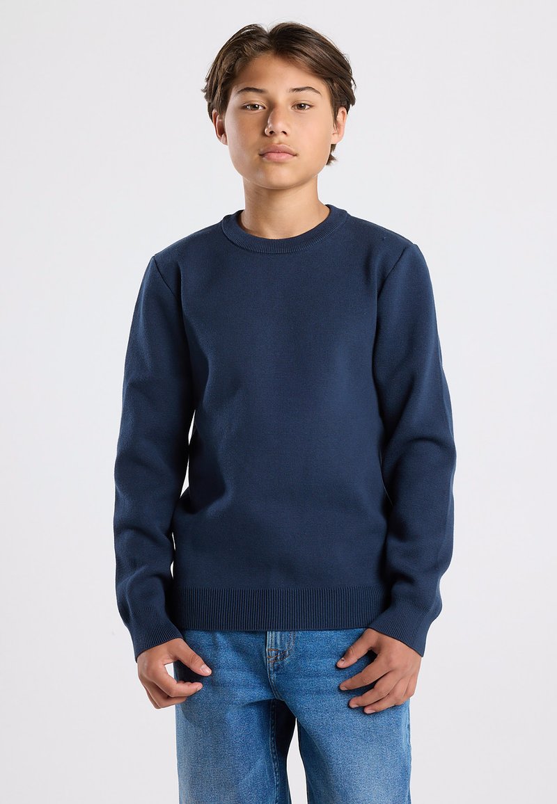 Tummansininen neulepaita, jossa on pyöreä kaula-aukko, pitkät hihat ja ribbaushelma. Yhdistetty vaaleansinisiin farkkuihin. Ei näkyviä kuvioita tai suunnittelua.