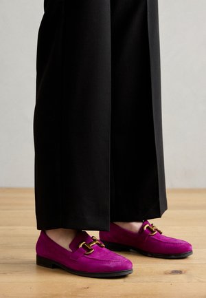 Fuksjowe zamszowe loafersy z akcentem w postaci złotej łańcuszka, w połączeniu z czarnymi spodniami, na drewnianej podłodze. Prosty, płaski design gumowej podeszwy.