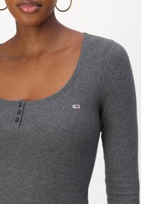 Top gri cu maneci lungi, cu dungi, și decolteu rotund, închidere cu trei nasturi și un logo brodat mic pe partea stângă a pieptului. Textură de țesătură netedă.