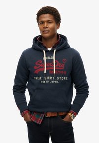 Marineblauwe hoodie met een kangoeroezak, trekkoord en rode grafische print. Gelaagd over een rood en groen geruit shirt. Draagt met donkere jeans en een horloge.