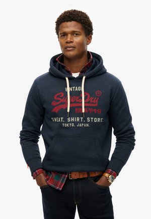 Marineblauwe hoodie met een kangoeroezak, trekkoord en rode grafische print. Gelaagd over een rood en groen geruit shirt. Draagt met donkere jeans en een horloge.