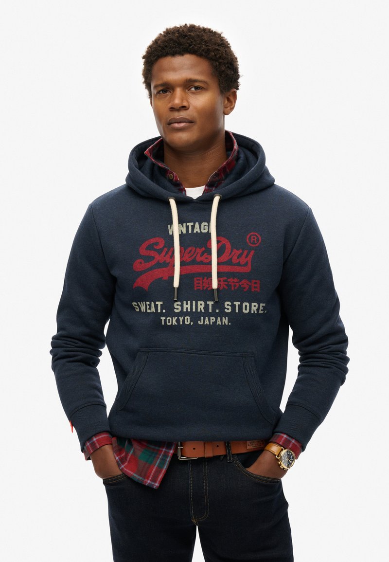 Marineblauwe hoodie met een kangoeroezak, trekkoord en rode grafische print. Gelaagd over een rood en groen geruit shirt. Draagt met donkere jeans en een horloge.