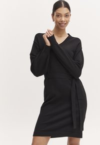 Abito nero in maglia con scollatura a V, maniche lunghe e una cintura coordinata. Presenta una silhouette aderente e una texture liscia.
