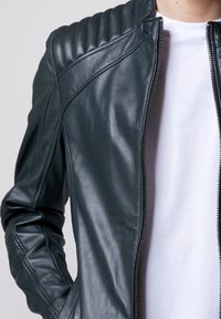 Giubbotto motociclistico in pelle verde scuro con dettagli trapuntati sulle spalle, zip frontale e design slim-fit, indossato sopra una maglietta bianca.