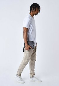 Borsa a tracolla nera con motivo, tenuta da una persona in maglietta bianca e pantaloni cargo beige, abbinata a sneaker bianche, in piedi contro uno sfondo bianco.
