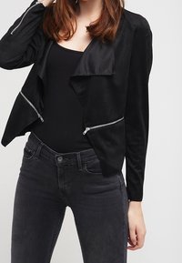 Veste en daim noir avec un devant drapé, des manches longues et des accents argentés zippés sur les côtés. Portée sur un haut noir et un jean.