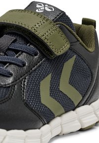 Svart och marinblå atletisk sko med mesh-tyg, ett olivgrönt rem och chevron-detaljer. Har en vit gummisula med segmenterat mönster.