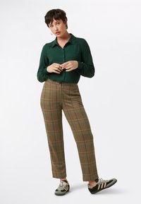 Groene blouse met stippen en knoopsluiting, gecombineerd met bruine geblokte broek. Schoenen hebben groene en zwarte details met veters.