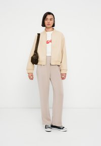 Μπεζ μπουφάν bomber, λευκή μπλούζα με κόκκινο κείμενο, μπεζ παντελόνια και μαύρα αθλητικά παπούτσια. Το ντύσιμο είναι casual και χαλαρό στο στυλ.