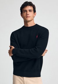 Schwarzer, gerippter Strickpullover mit Rundhalsausschnitt und langen Ärmeln. Verfügt über ein kleines rotes Logo auf der Brust. Eng anliegendes Design.